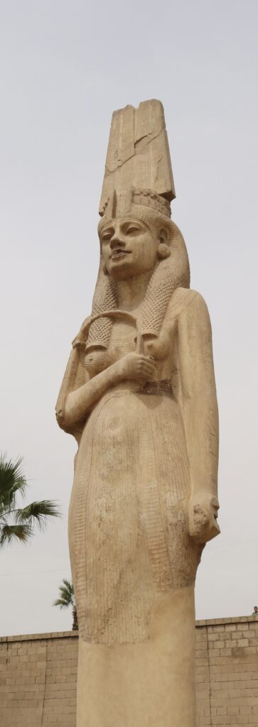 A tall sandstone statue of a queen with her hand on her chest.